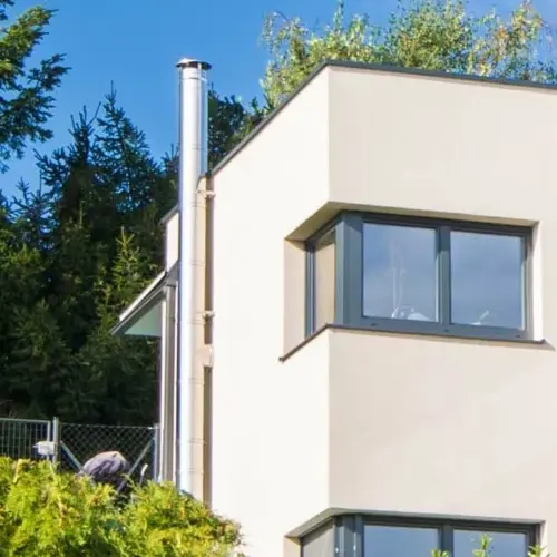 Moderner Edelstahlschornstein, der an der Seitenwand eines weiß gestrichenen Wohnhauses montiert ist, dass sich im Grünen befindet